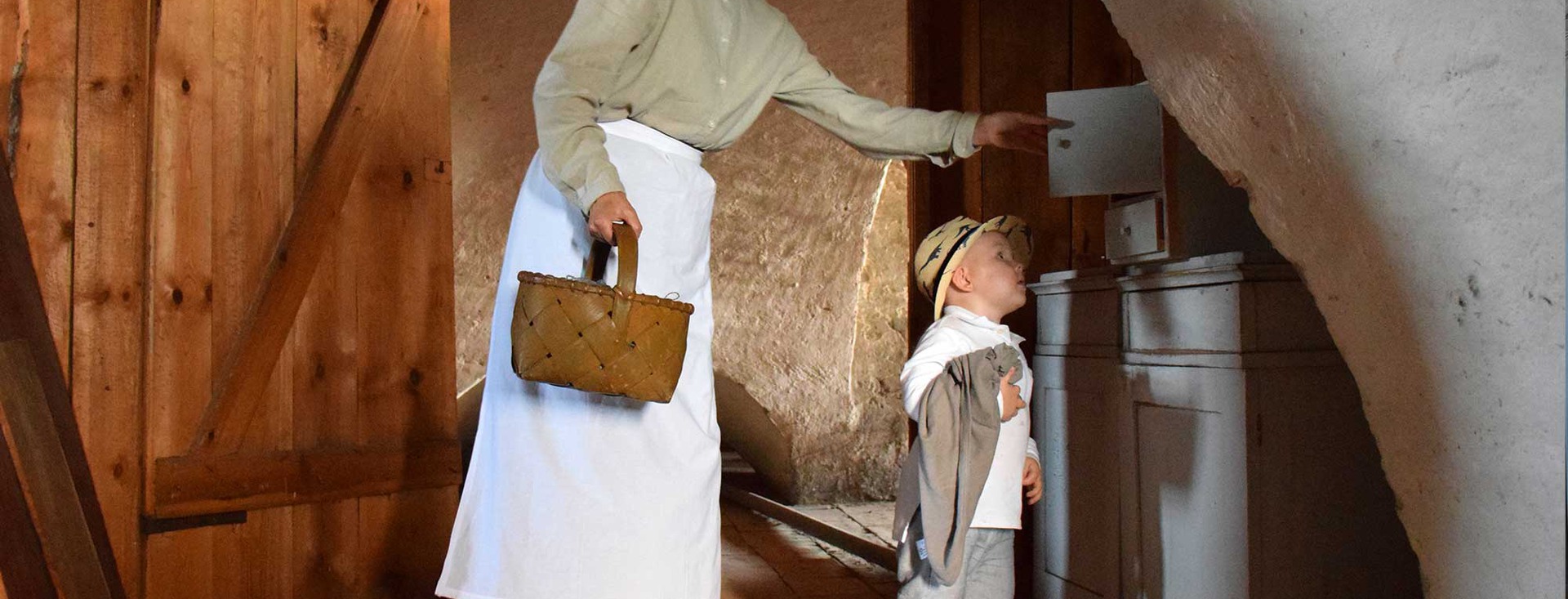 Ett litet barn iklädd stråhatt blir visad ett skåp av en traditionellt klädd person som håller en korg.